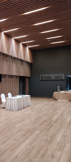 A spacious, modern hall with wooden flooring and walls featuring vertical wooden slats, illuminated by recessed lighting on the ceiling. A table draped with a white cloth has two chairs next to it, and there is a covered table with some catering equipment in the background.
