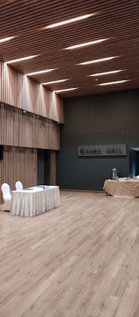 A spacious, modern hall with wooden flooring and walls featuring vertical wooden slats, illuminated by recessed lighting on the ceiling. A table draped with a white cloth has two chairs next to it, and there is a covered table with some catering equipment in the background.