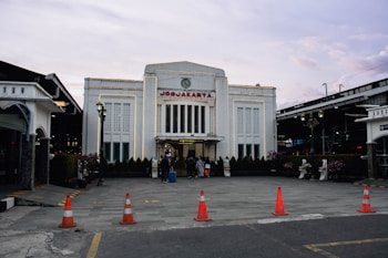 A large, white, art-deco-style building with the word 'Yogyakarta' displayed at the top is visible. The architecture includes vertical columns and intricate window designs. In the foreground, there are several orange traffic cones marking an area on the paved ground. People can be seen walking in and out of the building, and there are plants and lampposts enhancing the setting. The sky above is a blend of soft pastel colors, suggesting early morning or late afternoon.