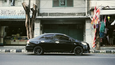 A black sedan car is parked on a street by the sidewalk in front of a row of older buildings. There are trees along the road, and one of them has several red and white flags tied to it. On the left, a shop with the partially visible sign 'BINTANG LOMB' can be seen.