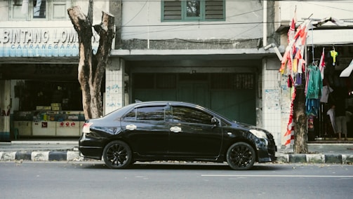 A black sedan car is parked on a street by the sidewalk in front of a row of older buildings. There are trees along the road, and one of them has several red and white flags tied to it. On the left, a shop with the partially visible sign 'BINTANG LOMB' can be seen.