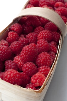 A basket filled with ripe red raspberries neatly arranged, with their textured surfaces visible. The basket is made of natural materials, adding a rustic feel.