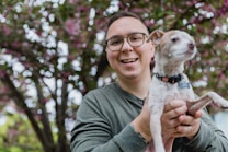 A person wearing glasses and a green shirt is holding a small dog with a pink collar, smiling at the camera. They are standing outdoors with a tree in full bloom in the background, featuring lush green leaves and pink flowers.