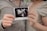 Two people holding a printed ultrasound image together, each using one hand. The ultrasound image is black and white, indicating a medical scan, possibly of a fetus. The individuals are wearing light-colored clothing, and one of them has a ring on their finger.