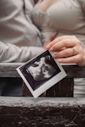 A close-up of hands holding an ultrasound image against a wooden background. The focus is on the photograph of the ultrasound, which is being gently grasped by a hand with manicured nails, showing a ring on one finger. The background and clothing suggest a casual setting.