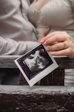 A close-up of hands holding an ultrasound image against a wooden background. The focus is on the photograph of the ultrasound, which is being gently grasped by a hand with manicured nails, showing a ring on one finger. The background and clothing suggest a casual setting.