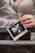 A close-up of hands holding an ultrasound image against a wooden background. The focus is on the photograph of the ultrasound, which is being gently grasped by a hand with manicured nails, showing a ring on one finger. The background and clothing suggest a casual setting.
