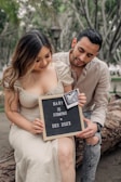 A couple is seated outdoors, smiling while holding a letter board with the message 'Baby is Coming Dec 2023' and an ultrasound photo attached. They appear happy and are showing genuine affection towards each other.