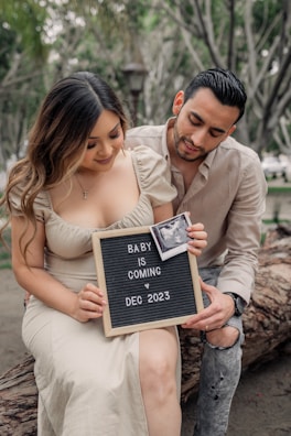 A couple is seated outdoors, smiling while holding a letter board with the message 'Baby is Coming Dec 2023' and an ultrasound photo attached. They appear happy and are showing genuine affection towards each other.