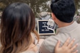 Two people are sitting by a body of water, focusing on a small sign that reads 'Baby is coming Dec 2023.' The sign is placed on the ground next to a pair of baby shoes and an ultrasound photo. The individuals appear to be a couple, with one person's hand resting on the other's back.