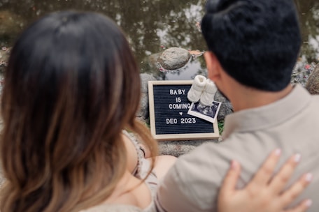 Two people are sitting by a body of water, focusing on a small sign that reads 'Baby is coming Dec 2023.' The sign is placed on the ground next to a pair of baby shoes and an ultrasound photo. The individuals appear to be a couple, with one person's hand resting on the other's back.