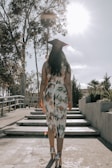 A graduate in a patterned dress and cap walks up a set of steps surrounded by trees and shrubbery under a bright sky with the sun shining.
