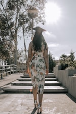 A graduate in a patterned dress and cap walks up a set of steps surrounded by trees and shrubbery under a bright sky with the sun shining.