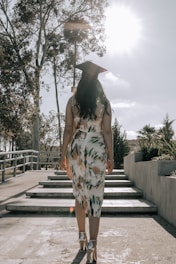 A graduate in a patterned dress and cap walks up a set of steps surrounded by trees and shrubbery under a bright sky with the sun shining.