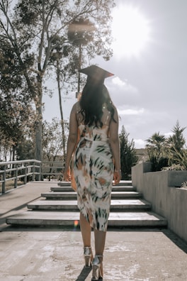 A graduate in a patterned dress and cap walks up a set of steps surrounded by trees and shrubbery under a bright sky with the sun shining.