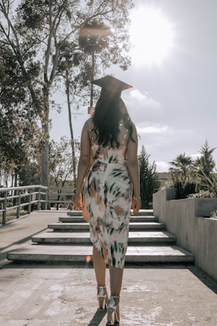 A graduate in a patterned dress and cap walks up a set of steps surrounded by trees and shrubbery under a bright sky with the sun shining.