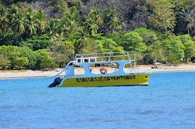 A yellow boat labeled 'SAMARA ADVENTURES' is floating on a blue body of water near a tropical shoreline lined with dense green palm trees and other vegetation.