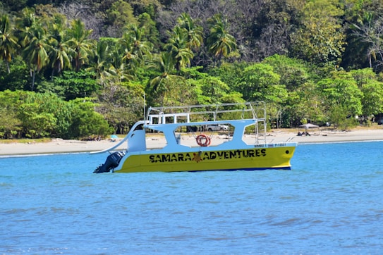 A yellow boat labeled 'SAMARA ADVENTURES' is floating on a blue body of water near a tropical shoreline lined with dense green palm trees and other vegetation.