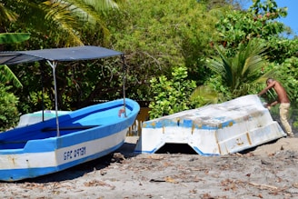 Two boats are positioned on a sandy beach surrounded by lush green foliage. One boat is painted blue and white with a canopy, while the other appears to be overturned. A person is leaning against the overturned boat, possibly making adjustments or repairs. The dense greenery in the background includes palm trees and other tropical plants, indicating a coastal environment.
