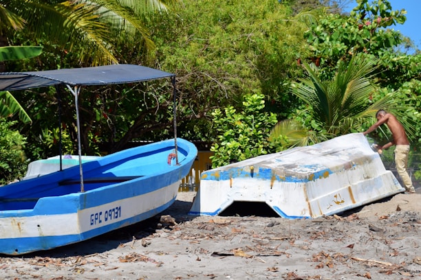 Two boats are positioned on a sandy beach surrounded by lush green foliage. One boat is painted blue and white with a canopy, while the other appears to be overturned. A person is leaning against the overturned boat, possibly making adjustments or repairs. The dense greenery in the background includes palm trees and other tropical plants, indicating a coastal environment.
