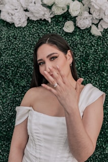 A woman in a white dress is showing off a ring on her hand. She is standing against a backdrop of green foliage and white flowers, and her expression appears to be joyful or proud.