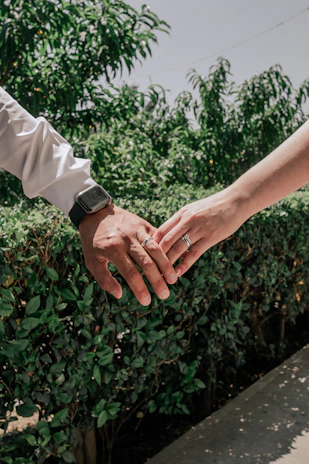 A close-up of intertwined hands with wedding bands against a lush green background.