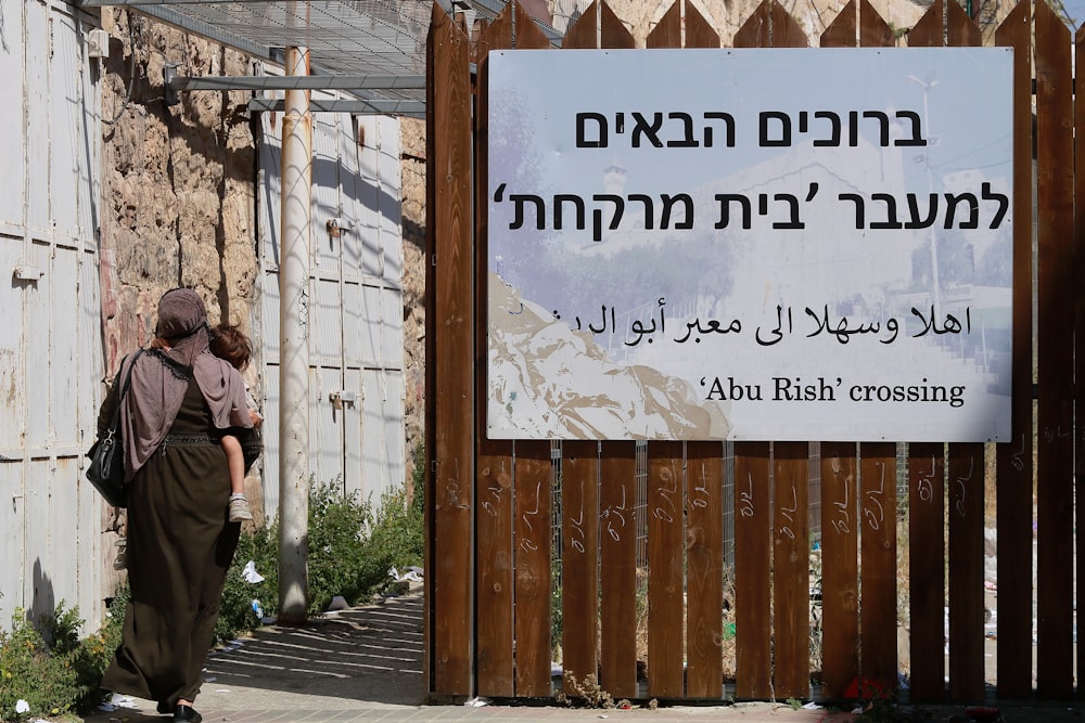 a woman walking past a wooden fence with a sign on it