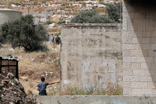 A young person is seen in a grassy area surrounded by dilapidated structures and a barbed wire fence. In the background, there are soldiers equipped with shields, partially hidden by trees. The setting appears to be rural with a mix of nature and man-made elements.