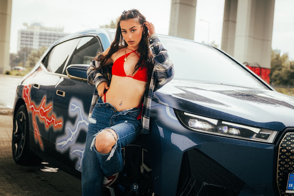 a woman in a red bra top leaning on a blue car