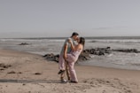 A couple embraces on a sandy beach with waves gently lapping against the shore. The man lifts the woman in a playful manner, and they gaze into each other's eyes, surrounded by rocks scattered along the water's edge.