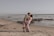 A couple embraces on a sandy beach with waves gently lapping against the shore. The man lifts the woman in a playful manner, and they gaze into each other's eyes, surrounded by rocks scattered along the water's edge.