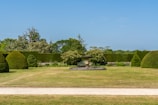 A well-manicured garden features a central stone urn on a pedestal surrounded by neatly trimmed hedges and bushes. The lawn is expansive, with a dirt path running across the foreground. In the background, various trees and shrubs enhance the greenery, and the sky is clear and blue.