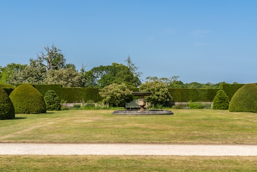 A well-manicured garden features a central stone urn on a pedestal surrounded by neatly trimmed hedges and bushes. The lawn is expansive, with a dirt path running across the foreground. In the background, various trees and shrubs enhance the greenery, and the sky is clear and blue.