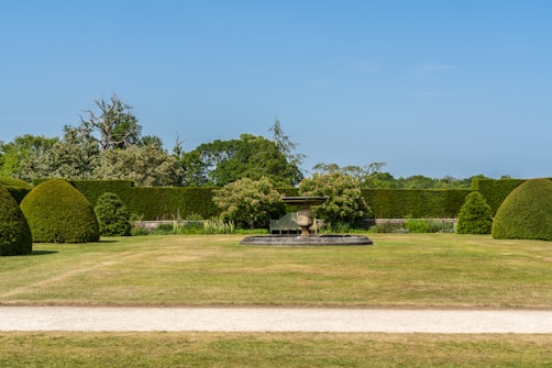 A well-manicured garden features a central stone urn on a pedestal surrounded by neatly trimmed hedges and bushes. The lawn is expansive, with a dirt path running across the foreground. In the background, various trees and shrubs enhance the greenery, and the sky is clear and blue.