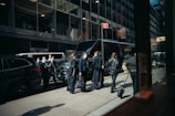 A group of people wearing business attire and face masks stand in front of a row of parked vehicles on a city street. They appear to be engaged in conversation or waiting. The background consists of tall office buildings with large windows.