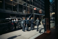 A group of people wearing business attire and face masks stand in front of a row of parked vehicles on a city street. They appear to be engaged in conversation or waiting. The background consists of tall office buildings with large windows.