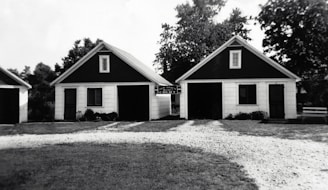 Two small, identical buildings with dark triangular roofs are positioned side by side. A gravel pathway leads up to them, bordered by grass. The buildings appear to have a rustic and simple architectural design, with white siding and black trim around the windows. Trees and other greenery are visible in the background.