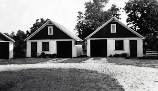 Two small, identical buildings with dark triangular roofs are positioned side by side. A gravel pathway leads up to them, bordered by grass. The buildings appear to have a rustic and simple architectural design, with white siding and black trim around the windows. Trees and other greenery are visible in the background.