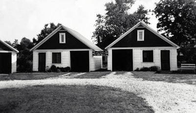 Two small, identical buildings with dark triangular roofs are positioned side by side. A gravel pathway leads up to them, bordered by grass. The buildings appear to have a rustic and simple architectural design, with white siding and black trim around the windows. Trees and other greenery are visible in the background.