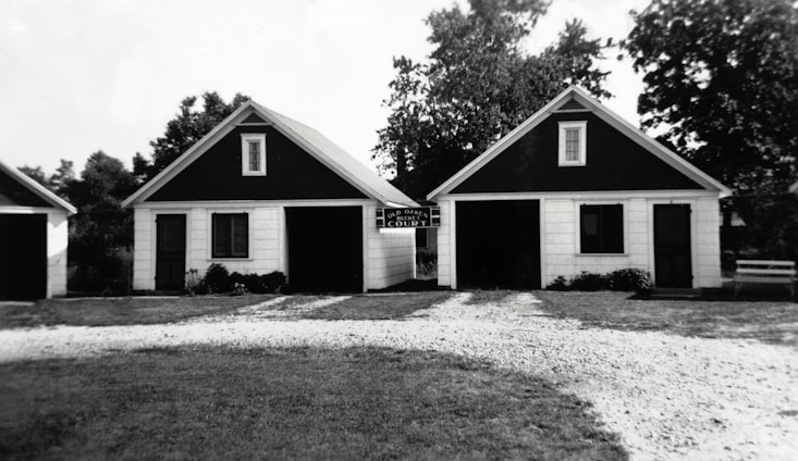 Two small, identical buildings with dark triangular roofs are positioned side by side. A gravel pathway leads up to them, bordered by grass. The buildings appear to have a rustic and simple architectural design, with white siding and black trim around the windows. Trees and other greenery are visible in the background.
