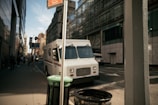 A white delivery truck is parked on an urban street near a modern glass building. The street is lined with tall buildings and a few pedestrians are walking on the sidewalk. A trash bin and a pole are in the foreground.
