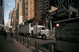 A large semi-truck is navigating a street surrounded by tall, modern buildings. An NYU Langone Health ambulance is parked nearby, and a few pedestrians are walking along the sidewalk. Metal bollards are lined up along the edge of the sidewalk. The overall atmosphere seems busy and urban.