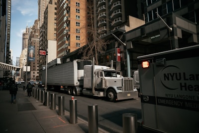 A large semi-truck is navigating a street surrounded by tall, modern buildings. An NYU Langone Health ambulance is parked nearby, and a few pedestrians are walking along the sidewalk. Metal bollards are lined up along the edge of the sidewalk. The overall atmosphere seems busy and urban.
