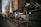 A large semi-truck is navigating a street surrounded by tall, modern buildings. An NYU Langone Health ambulance is parked nearby, and a few pedestrians are walking along the sidewalk. Metal bollards are lined up along the edge of the sidewalk. The overall atmosphere seems busy and urban.