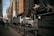 A large semi-truck is navigating a street surrounded by tall, modern buildings. An NYU Langone Health ambulance is parked nearby, and a few pedestrians are walking along the sidewalk. Metal bollards are lined up along the edge of the sidewalk. The overall atmosphere seems busy and urban.