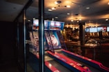 An indoor amusement area features an arcade game with several lanes for skee-ball. The game is housed in a glass enclosure, and the lanes are vibrant with pink and blue accents. Overhead lighting casts a warm glow, highlighting the modern and slightly retro atmosphere. The background includes other games or machines, giving the space a lively ambiance.