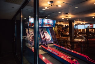 An indoor amusement area features an arcade game with several lanes for skee-ball. The game is housed in a glass enclosure, and the lanes are vibrant with pink and blue accents. Overhead lighting casts a warm glow, highlighting the modern and slightly retro atmosphere. The background includes other games or machines, giving the space a lively ambiance.