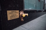 A metallic standpipe and sprinkler connection is mounted on a dark exterior wall, accompanied by a sign indicating the combination standpipe and sprinkler system. The sign mentions that all floors except certain numbered ones are fully sprinklered. The sidewalk next to the wall is tiled, and a large glass window reflects the urban environment.