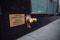 A metallic standpipe and sprinkler connection is mounted on a dark exterior wall, accompanied by a sign indicating the combination standpipe and sprinkler system. The sign mentions that all floors except certain numbered ones are fully sprinklered. The sidewalk next to the wall is tiled, and a large glass window reflects the urban environment.