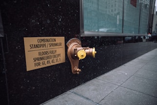 A metallic standpipe and sprinkler connection is mounted on a dark exterior wall, accompanied by a sign indicating the combination standpipe and sprinkler system. The sign mentions that all floors except certain numbered ones are fully sprinklered. The sidewalk next to the wall is tiled, and a large glass window reflects the urban environment.